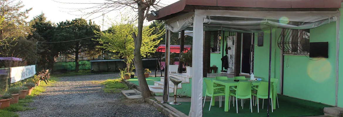 Yeşil boyalı bir binanın önünde masa ve sandalyelerle düzenlenmiş açık oturma alanı, çakıllı yol ve ağaçlar.